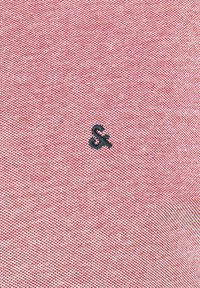 Rode textuurstof met een subtiele weving; heeft een klein zwart geborduurd logo in het midden onderaan.