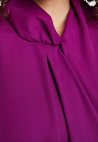 Amethystfarbene Bluse mit Schal-Ausschnitt und gefaltetem Detail, gefertigt aus weichem, glattem Material, das ein fließendes und elegantes Design zeigt.