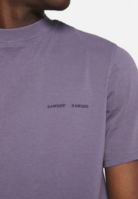 Gros plan sur une personne portant un t-shirt crew-neck violet avec le texte noir "SAMSOE SAMSOE" sur la poitrine gauche.
