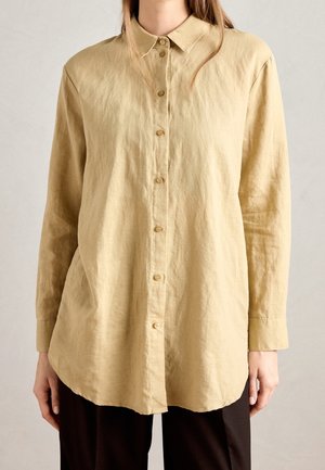 Femme portant une chemise beige ample à manches longues avec col et boutons, associée à un pantalon foncé, debout devant un fond uni.