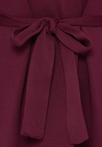 Gros plan d’un tissu tricot bordeaux noué en un nœud plat avec deux larges boucles et deux extrémités pendantes.
