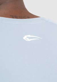 Camiseta ajustada de color azul claro con cuello redondo. Presenta un pequeño logotipo blanco en la parte posterior y una tela suave y texturizada.