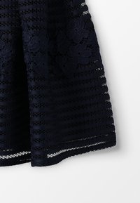 Tummansininen pitsihame vaakasuoralla raidoituksella, jossa on kukkakirjontaa ja läpinäkyviä verkko-osiota helman kohdalla.