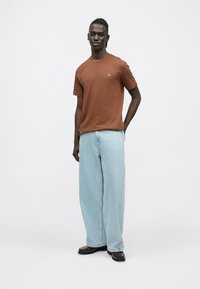 Camiseta marrón de manga corta, corte holgado, combinada con vaqueros de pierna ancha en azul claro. La modelo está de pie con las manos en los bolsillos, llevando zapatos negros.