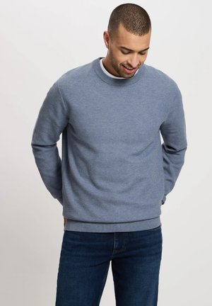 Niebieska dzianinowa bluza z okrągłym dekoltem i ściągaczami u rękawów, noszona na białej koszulce, w połączeniu z ciemnoniebieskimi dżinsami. Gładka tekstura.