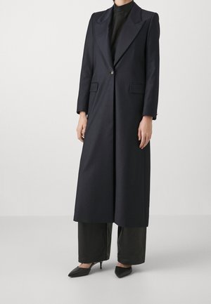 Femme portant un long manteau noir sur mesure par-dessus un pantalon à jambes larges et des talons noirs à bouts pointus, debout devant un fond clair uni.