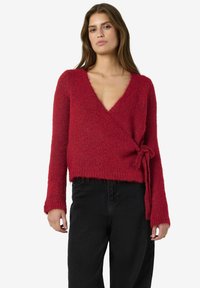 Kvinna iklädd en röd fluffig omslagströja med långa ärmar knuten i midjan och svarta höga byxor, stående mot en vit bakgrund.