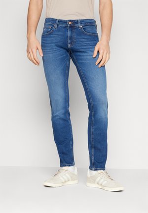 Jeans de mezclilla azul oscuro con corte slim, con un ligero desgaste, diseño clásico de cinco bolsillos y cierre con botón de metal, combinados con zapatillas de color claro.