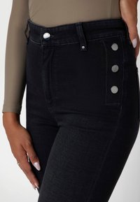 High-waisted black jeans with a smooth texture, featuring silver button accents on the side and a classic five-pocket design.