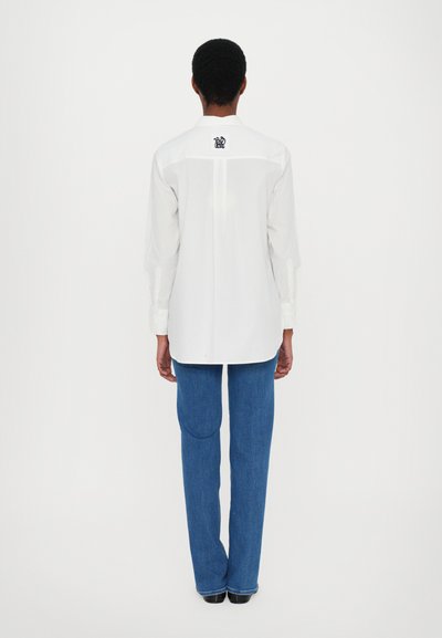 Camicia bianca a maniche lunghe con yoke posteriore e logo ricamato, abbinata a jeans in denim blu con dettagli sulle tasche.