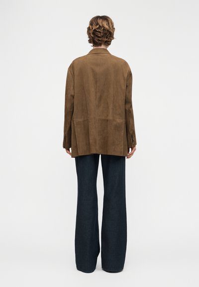 Personne aux cheveux courts et bouclés portant une veste ample en daim marron et un pantalon large bleu foncé, debout de dos sur un fond blanc.