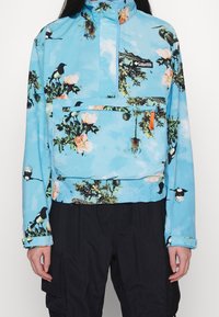 Light blue pullover jacket with floral and bird print, front zip pocket, and black pants on a standing person.