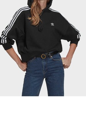 Vrouw draagt een zwarte Adidas hoodie met witte strepen en logo, gecombineerd met een blauwe spijkerbroek en een zwarte riem met gouden gesp.