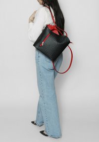 Borsa nera con texture, rifiniture rosse, tasca con zip e tracolla regolabile, indossata sulla spalla con jeans leggeri color blu.