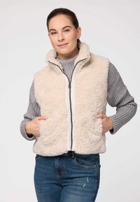 Kratkā krēma krāsas mākslīgās kažokādas veste ar augstu apkakli, rāvējslēdzēju un divām priekšējām kabatām. Valkāta virs pelēka adīta džempera un ziliem džinsiem.