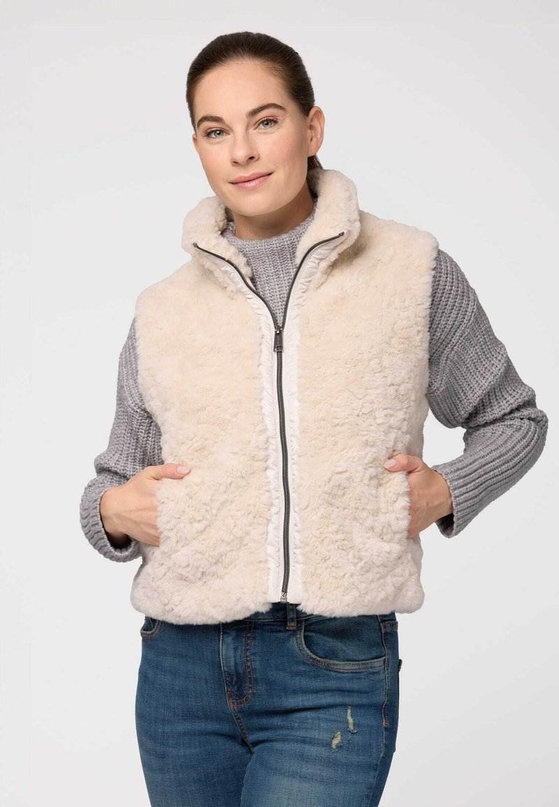 Kratkā krēma krāsas mākslīgās kažokādas veste ar augstu apkakli, rāvējslēdzēju un divām priekšējām kabatām. Valkāta virs pelēka adīta džempera un ziliem džinsiem.