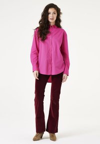 Camisa magenta de botones con un corte holgado y mangas largas, combinada con pantalones de pana burdeos ajustados y botas de tacón beige.