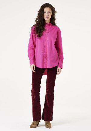 Magenta Hemdbluse mit lockerem Schnitt und langen Ärmeln, kombiniert mit figurbetonten bordeauxrotenCorduroy-Bootcut-Hosen und beigen Stiefeletten mit Absatz.