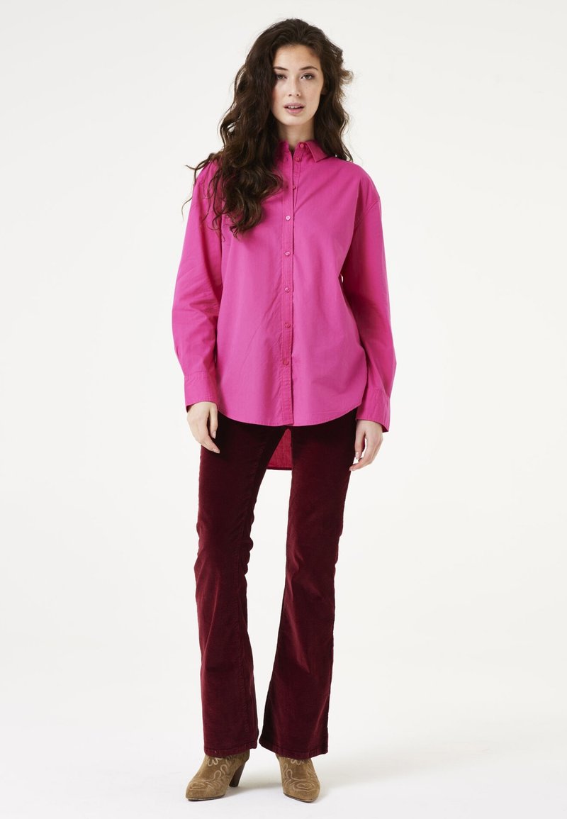 Camisa magenta de botones con un corte holgado y mangas largas, combinada con pantalones de pana burdeos ajustados y botas de tacón beige.
