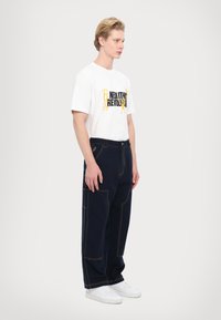 White t-shirt with black and yellow graphic text, paired with dark blue loose-fitting cargo pants featuring contrasting stitching and pockets.