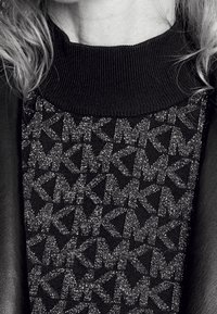 Maglione nero con un colletto alto a coste e un motivo scintillante che presenta design di lettere intrecciate. Accentuato con un tessuto nero liscio.