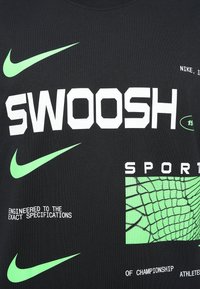 Črna majica s kratkimi rokavi, ki prikazuje velik bel napis "SWOOSH" in "SPORTS", z zelenim Nike swoosh logotipom in grafiko mrežnega vzorca.
