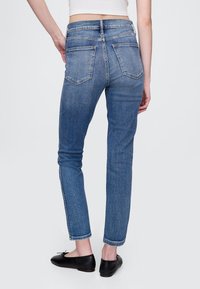 Mėlynos spalvos liesi džinsai iš džinsinio audinio, turintys vidutinio aukščio liemenį, su galinėmis kišenėmis ir minimaliais siuvimo elementais, derinami su juodomis lakuotomis bateliais.