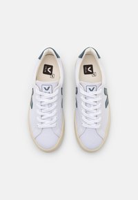 Tênis de lona brancos com detalhes em cinza claro, apresentando bico arredondado, design com atacadores e sola de borracha. Logótipo visível na lingueta.