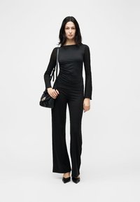 Ženska stoji v črni oprijeti majici z dolgimi rokavi in širokih hlačah z visokim pasom, v roki drži črno torbico in nosi črne koničaste čevlje na belem ozadju.