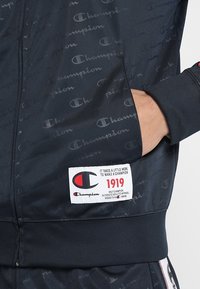 Mörkblå atletisk jacka med ett upprepande logomönster. Har en dragkedjeficka och en märkt etikett på sidan. Slät struktur, avslappnad passform.