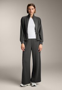 Šedá rebrovaná bunda so zipsom a s detaily šnúrky v kombinácii s voľnými šedými nohavicami. Pod ňou biele priliehavé tričko. Neformálne športové oblečenie.