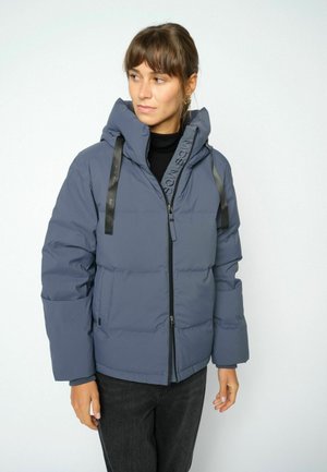 Donna che indossa una giacca imbottita blu con collo alto e zip nera, che guarda a sinistra su uno sfondo bianco semplice.