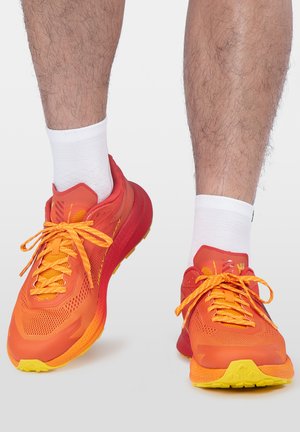 Par de zapatillas de correr naranjas con suelas y cordones amarillos, usadas con calcetines blancos hasta el tobillo en piernas peludas, sobre un fondo liso.