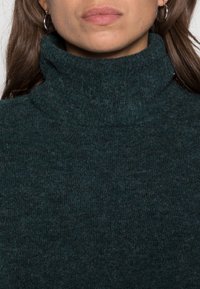 Maglione a collo alto di colore verde scuro, realizzato in tessuto a maglia testurizzato. Presenta un colletto alto e una vestibilità aderente, adatto per essere indossato a strati e per il clima più freddo.