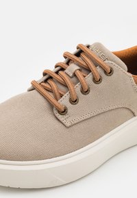 Zapatillas de lona beige con cordones marrones, ojales dorados, tejido texturizado y suela de goma blanca, que cuentan con una punta redondeada y un diseño minimalista.