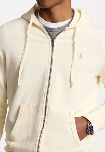 Creme zip-up hoodie med en glat overflade, frontlommer, snoretræk og et lille logo på venstre bryst. Båret over en grå skjorte.
