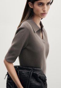 Donna con capelli lunghi e scuri che indossa una maglia a maniche corte di colore taupe, tenendo una grande borsa intrecciata in pelle nera, su uno sfondo semplice.