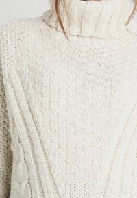 Sweter w kolorze kremowym, z dzianiny, z wysokim prążkowanym kołnierzem, z teksturowanym wzorem w warkocze z przodu oraz rękawami raglanowymi.