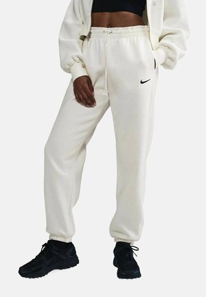 Personne portant un jogging Nike écru avec cordon, un crop top noir, une veste crème zippée et des chaussures de sport noires, debout sur fond blanc.
