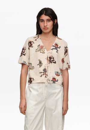 Mujer joven que lleva un top de punto de manga corta beige con estampados abstractos de animales en colores y lazo delantero, combinado con pantalones blancos holgados.