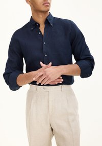 Camicia di lino navy con colletto button-down, maniche lunghe arrotolate ai polsi, abbinata a pantaloni beige chiaro su misura con pieghe frontali.