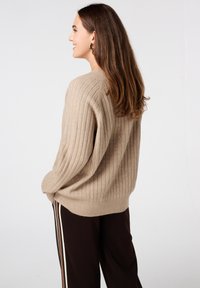 Pull en maille beige à côtes avec des manches longues et une coupe décontractée, associé à un pantalon sombre avec des rayures sur les côtés. Texture lisse, design simple.