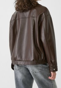 Chaqueta marrón de cuero sintético con un corte relajado, dobladillo acanalado y puños con botones, combinada con vaqueros gris claro. Textura suave y brillante.