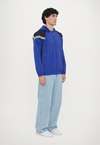 Sininen kevyt takki, jossa on musta-valkoiset raidalliset olkaimet, vetoketjulliset taskut ja huppu. Slim fit -malli, jossa on elastiset hihansuut.