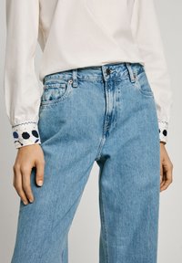 Ljusblå jeans med hög midja, med framfickor och knappstängning. Vit topp med dekorativa broderade manschetter.