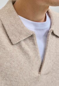 Beige strikpullover med flad krave, der har en struktureret overflade og en dyb V-hals. En hvid T-shirt med rund hals er synlig under.