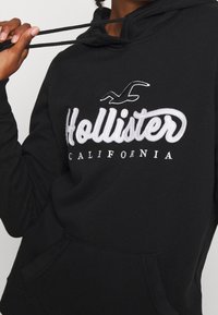 Felpa nera con cappuccio realizzata in materiale morbido, con un grande logo ricamato bianco "Hollister" e una piccola grafica di gabbiano; presenta una tasca frontale e cordini.