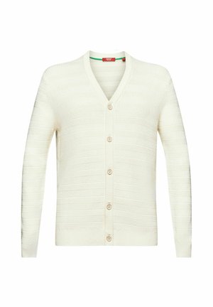 Mann trägt weißen Strickcardigan und rotes Hemd, hält eine marineblaue Umhängetasche am Gurt quer vor der Brust.