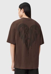 Brun bomulds-t-shirt med stort sort hjertegrafik med drypseffekt på ryggen. Korte ærmer og afslappet pasform.