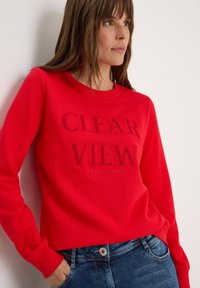 Sweatshirt rouge avec col rond et manches longues, présentant un graphique en relief avec le texte "CLAIR REGARD RÉFLEXION" dans une couleur contrastante. Assorti avec un jean bleu.